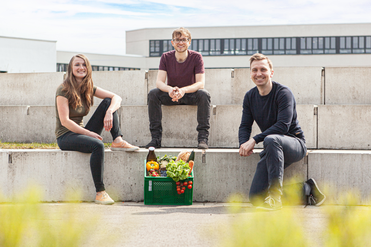 Ein Bild der Gründer. Dazwischen steht eine Gemüsekiste.